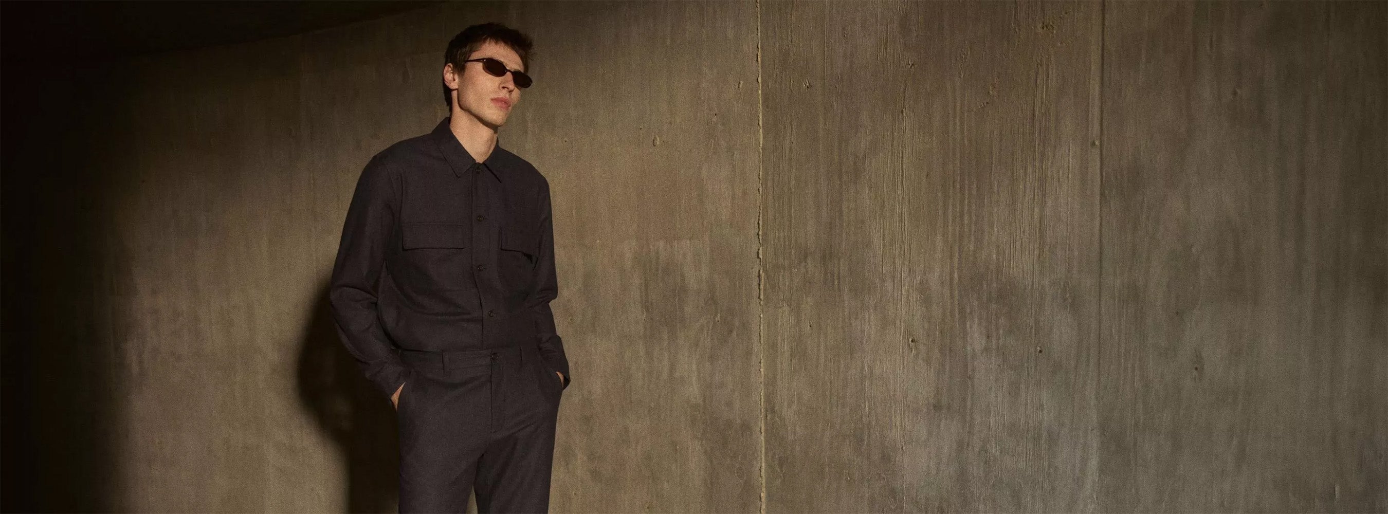 Männliches Model mit dunkler Sonnenbrille präsentiert ein monochromes, dunkelgraues Outfit bestehend aus einem Hemd mit Brusttaschen und einer passenden Hose vor einer rauen Betonwand mit markantem Schattenwurf.