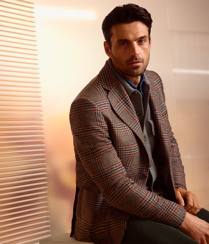 Ein Model trägt einen mehrfarbigen Blazer mit Karomuster in Braun und Rot über einer grauen Strickjacke und einem blauen Hemd. Dazu kombiniert er eine dunkle Hose. Das Porträt zeigt ihn sitzend vor einem hellen Hintergrund mit horizontalen Lamellen.