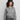 Model mit markantem schwarzem Hut präsentiert eine hellgraue Zip-Strickjacke von Extreme Cashmere über einem grauen Shirt, kombiniert mit dunkelgrauem Unterteil vor hellem Hintergrund.