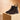 Paar dunkelbraune Chelsea Boots von Doucal’s aus Veloursleder mit elastischem Einsatz und gelber Zuglasche, auf einem hellen Podest vor warmem Jalousie-Hintergrund.