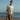 Ein Ganzkörperfoto eines männlichen Models, das am Strand vor einem ruhigen Meer und klarem blauen Himmel steht. Er trägt ein strukturiertes, cremefarbenes (off-white) kurzärmeliges Poloshirt aus Feinstrick mit offenem Stichmuster. Dazu kombiniert er eine hellbraune Leinenhose und hält eine segeltuchfarbene Tragetasche.