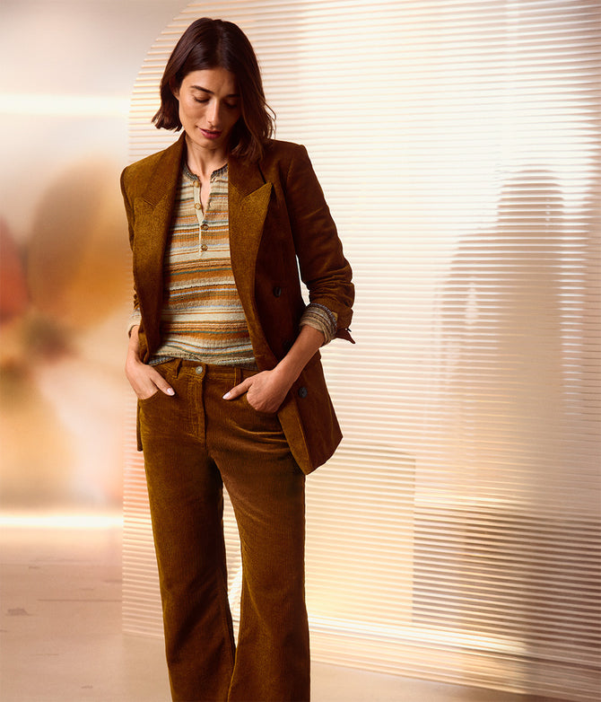 Ein Model trägt einen braunen Cordblazer mit passender, weit geschnittener Cordhose und einem gestreiften Strickshirt in Beige, Braun und Senfgelb von Isabel Marant. Der Blazer ist lässig offen getragen, die Ärmel sind hochgekrempelt. Die Hände stecken in den Hosentaschen. Im Hintergrund ist eine helle, modern gestaltete Wand mit horizontalen Lamellen zu sehen.