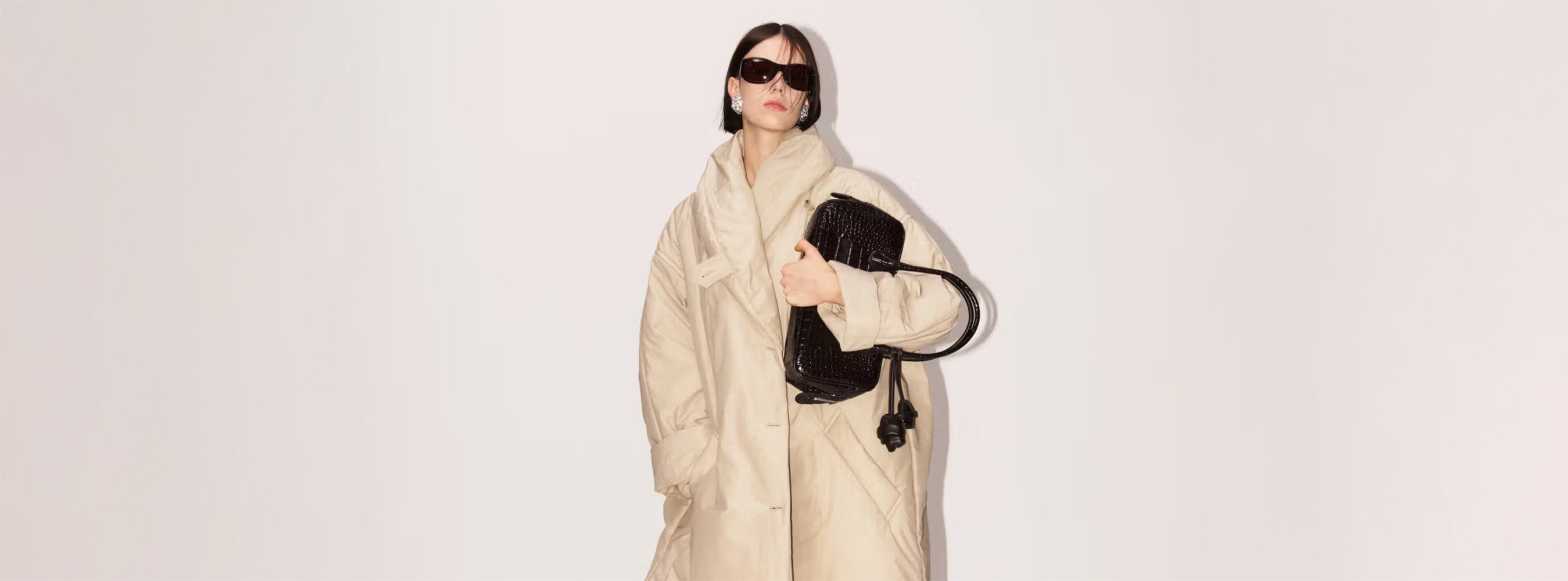 Model präsentiert einen voluminösen, beigefarbenen Oversize-Mantel, kombiniert mit einer dunklen Sonnenbrille, großen Ohrringen und einer schwarzen, strukturierten Handtasche von Magda Butrym vor hellem Hintergrund.